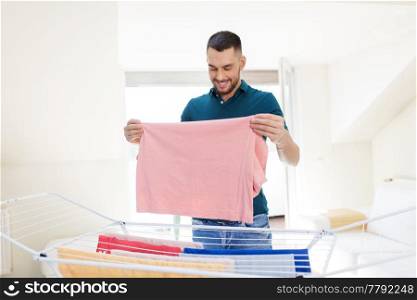 laundry and household concept - happy man taking clothes from drying rack and putting them to basket at home. smiling man with laundry and drying rack at home