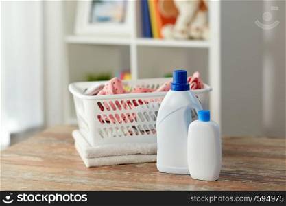 laundry, wash and housekeeping concept - baby clothes in basket with detergent and conditioner bottles on wooden table at home. baby clothes in laundry basket with detergent