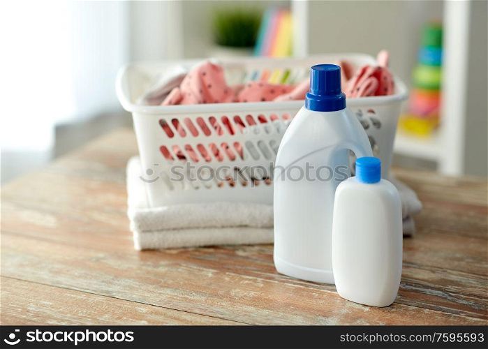 laundry, wash and housekeeping concept - baby clothes in basket with detergent and conditioner bottles on wooden table at home. baby clothes in laundry basket with detergent