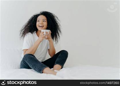 Lazy morning and bedtime concept. Positive Afro woman holds mug of hot tea or coffee, sits in bed, looks gladfully aside, enjoys good weather, wears comfortable clothes, white wall in background