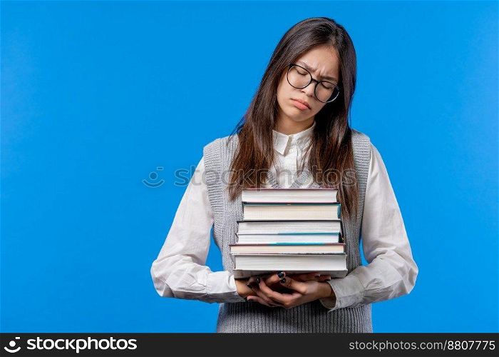 Lazy schoolgirl or student is dissatisfied with amount of books homework on blue background. Girl with long hair in displeasure, she is annoyed, discouraged frustrated by studies. High quality photo. Lazy schoolgirl or student is dissatisfied with amount of books homework on blue background. Girl with long hair in displeasure, she is annoyed, discouraged frustrated by studies