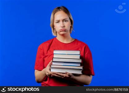 Lazy schoolgirl or student is dissatisfied with amount of books homework on blue background. Girl in displeasure, she is annoyed, discouraged frustrated by studies. High quality photo. Lazy schoolgirl or student is dissatisfied with amount of books homework on blue
