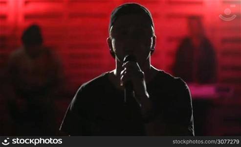Lead singer with his eyes closed holding microphone and singing song, blurred band members in background. Young passionate vocalist performing song for his adoring fans. Musicians playing musical instruments and singing songs in the club