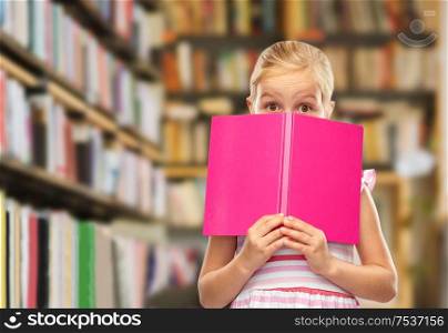 learning, education and school concept - little girl hiding behind book over library shelves background. little girl hiding over book at library