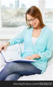 leasure and home concept - smiling woman in eyeglasses reading book and sitting on couch at home