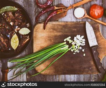 Leaves of green onion on a kitchen board with a knife, near a frying pan with fried pork ribs