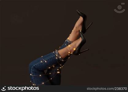 Legs of a woman in elegant black high-heeled shoes with bright christmas lights on a black background. As the layout for your ideas. Legs of a woman in jeans and shoes decorated with yellow garlands around a black background