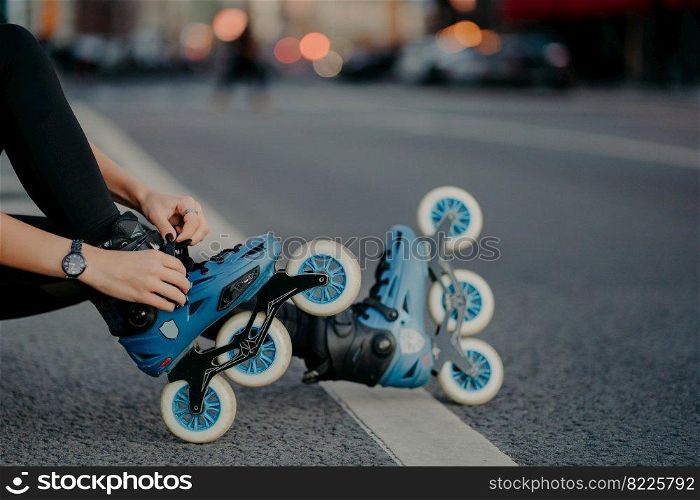 Legs of unrecognizable woman puts on rollerskates going to have exercises outdoor leads active lifestyle poses on street road prepares for inline skating. Faceless skater goes in for dangerous sport