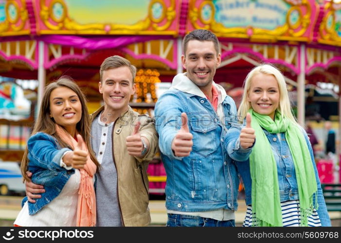 leisure, amusement park and friendship concept - group of smiling friends showing thumbs up with carousel on the back