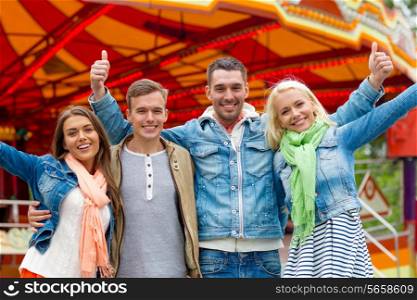 leisure, amusement park and friendship concept - group of smiling friends showing thumbs up with carousel on the back