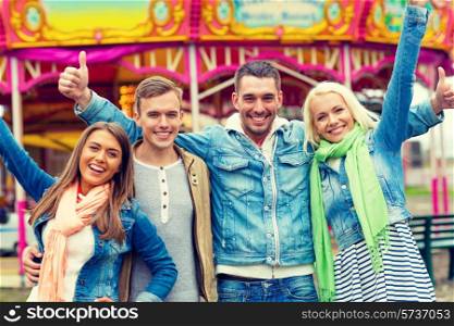 leisure, amusement park and friendship concept - group of smiling friends showing thumbs up with carousel on the back