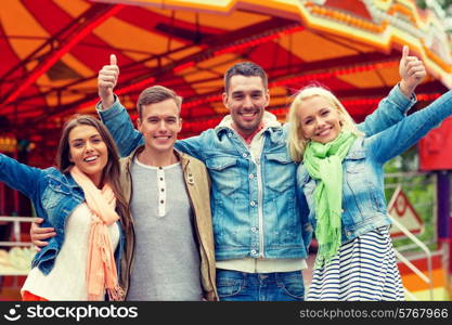 leisure, amusement park and friendship concept - group of smiling friends showing thumbs up with carousel on the back