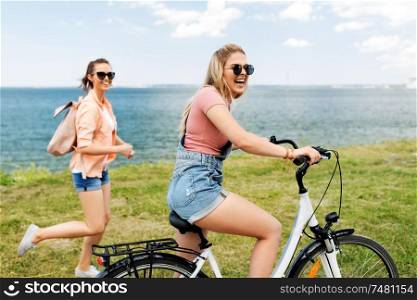 leisure and friendship concept - happy smiling teenage girl riding bicycle and her friend running nearby at seaside in summer. teenage girls or friends with bicycle in summer