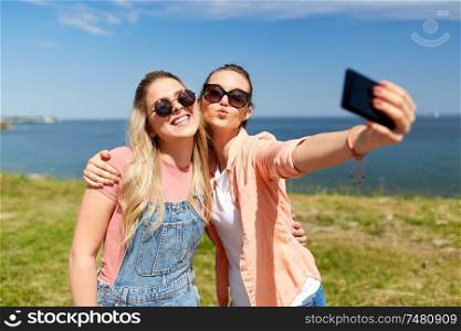 leisure and friendship concept - happy smiling teenage girls or best friends in sunglasses hugging and taking selfie by smartphone at seaside in summer. teenage girls or friends taking selfie in summer