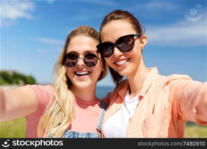 leisure and friendship concept - happy smiling teenage girls or best friends in sunglasses hugging and taking selfie at seaside in summer. teenage girls or friends taking selfie in summer