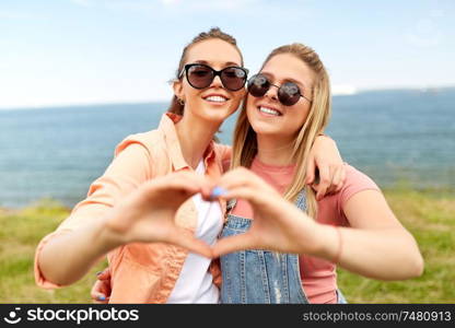 leisure and friendship concept - happy smiling teenage girls or best friends in sunglasses at seaside in summer hugging and making hand heart gesture. teenage girls or best friends at seaside in summer