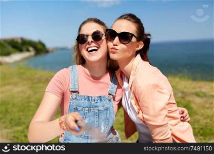 leisure and friendship concept - happy smiling teenage girls or best friends in sunglasses hugging and taking picture by selfie stick at seaside in summer. teenage girls or friends taking selfie in summer