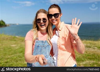 leisure and friendship concept - happy smiling teenage girls or best friends in sunglasses waving hand and taking picture by selfie stick at seaside in summer. teenage girls or friends taking selfie in summer