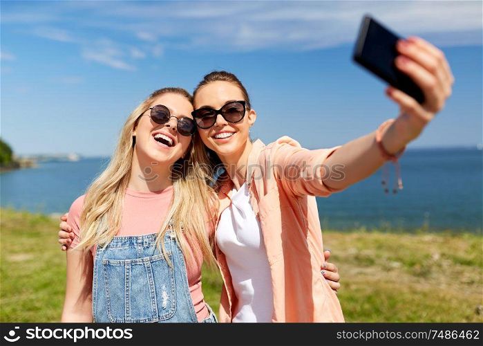 leisure and friendship concept - happy smiling teenage girls or best friends in sunglasses hugging and taking selfie by smartphone at seaside in summer. teenage girls or friends taking selfie in summer