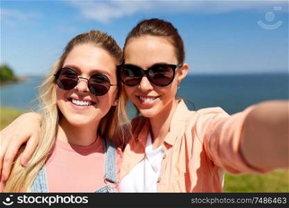 leisure and friendship concept - happy smiling teenage girls or best friends in sunglasses hugging and taking selfie at seaside in summer. teenage girls or friends taking selfie in summer