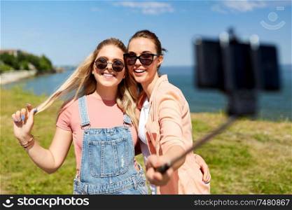 leisure and friendship concept - happy smiling teenage girls or friends in sunglasses hugging and taking picture by smartphone on selfie stick at seaside in summer. teenage girls or friends taking selfie in summer
