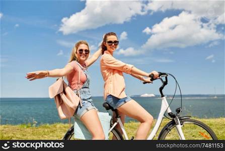 leisure and friendship concept - happy smiling teenage girls or friends riding bicycle together at seaside in summer. teenage girls or friends riding bicycle in summer
