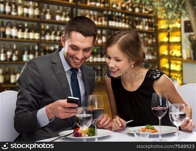 leisure and luxury concept - smiling couple with smartphone and food over restaurant or wine bar background. smiling couple eating main course at restaurant