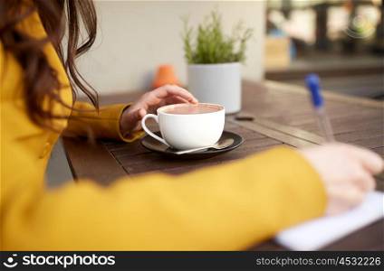 leisure and people concept - close up of young woman or teenage girl with cup drinking cocoa and writing to notebook at city street cafe terrace. close up of woman drinking cocoa at cafe