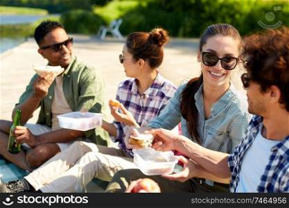 leisure and people concept - group of happy friends having picnic and eating sandwiches on lake pier in summer. happy friends having picnic on lake pier in summer