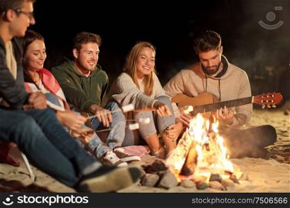 leisure and people concept - group of smiling friends sitting at camp fire on beach, roasting marshmallow and playing guitar at night. friends roasting marshmallow and playing guitar