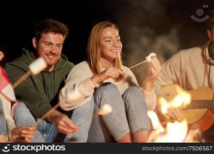 leisure and people concept - group of smiling friends sitting at camp fire, roasting marshmallow and playing guitar at night. friends roasting marshmallow and playing guitar