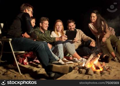 leisure and people concept - group of smiling friends sitting at camp fire with tablet pc computer on beach at night. friends with tablet pc at night camp fire on beach