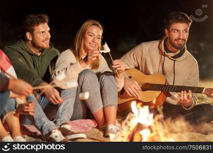 leisure and people concept - group of smiling friends sitting at camp fire on beach, roasting marshmallow and playing guitar at night. friends roasting marshmallow and playing guitar