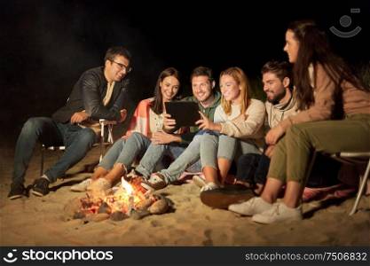leisure and people concept - group of smiling friends sitting at camp fire with tablet pc computer on beach at night. friends with tablet pc at fire on beach at night