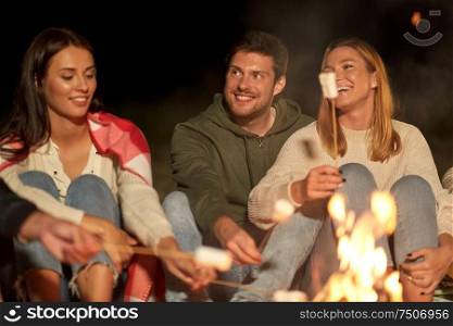 leisure and people concept - group of smiling friends sitting at camp fire and roasting marshmallow at night. friends roasting marshmallow on camp fire at night