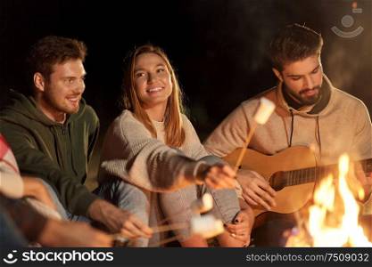 leisure and people concept - group of smiling friends sitting at camp fire, roasting marshmallow and playing guitar at night. friends roasting marshmallow and playing guitar