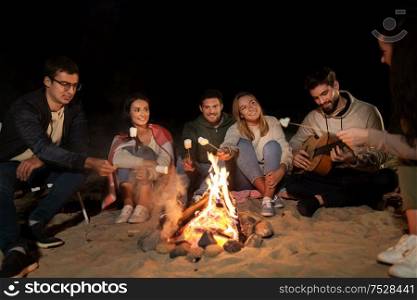 leisure and people concept - group of smiling friends sitting at camp fire on beach and roasting marshmallow at night. friends roasting marshmallow on camp fire on beach