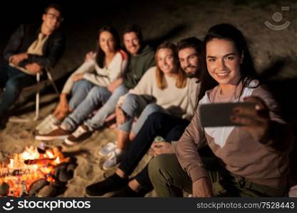 leisure and people concept - group of smiling friends sitting at camp fire taking selfie by smartphone on beach at night. happy friends taking selfie at camp fire on beach