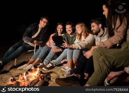leisure and people concept - group of smiling friends sitting at camp fire with tablet pc computer on beach at night. friends with tablet pc at fire on beach at night
