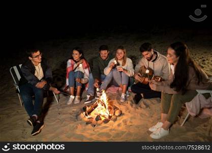 leisure and people concept - group of smiling friends sitting at camp fire on beach and roasting marshmallow at night. friends roasting marshmallow on camp fire on beach
