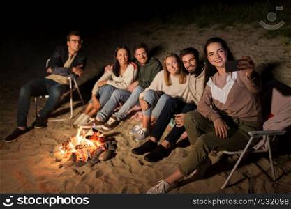 leisure and people concept - group of smiling friends sitting at camp fire taking selfie by smartphone on beach at night. happy friends taking selfie at camp fire on beach
