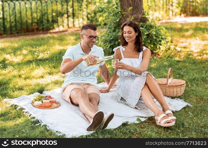 leisure and people concept - happy couple having picnic and pouring wine to glass at summer park. happy couple with wine having picnic at park