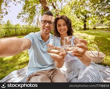 leisure and people concept - happy couple with drinks having picnic at summer park. couple drinking wine and taking selfie at park