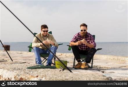 leisure and people concept - happy friends fishing and eating sandwiches on pier at sea. happy friends fishing and eating sandwiches
