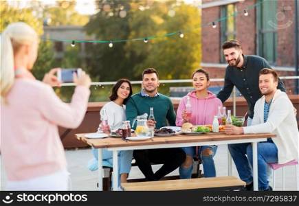 leisure and people concept - happy friends with non alcoholic drinks having dinner party on rooftop in summer and photographing by smartphone. happy friends photographing at rooftop party