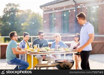 leisure and people concept - happy man grilling meat on bbq and friends at rooftop party. man grilling meat on bbq at rooftop party
