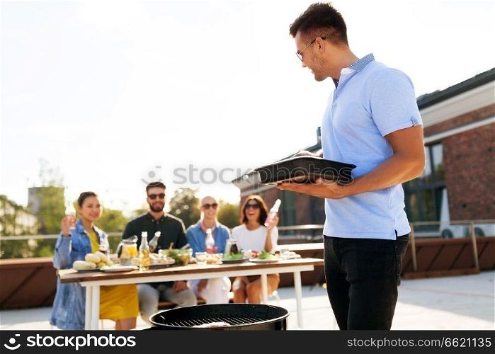 leisure and people concept - happy man with tray cooking on bbq and friends at rooftop party. man cooking on bbq and friends at rooftop party
