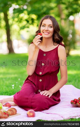 leisure and people concept - happy smiling woman eating strawberry sitting on picnic blanket at summer park. happy woman eating strawberry on picnic at park