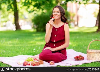 leisure and people concept - happy smiling woman eating strawberry sitting on picnic blanket at summer park. happy woman eating strawberry on picnic at park