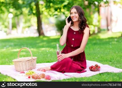 leisure and people concept - happy smiling woman with fizzy drink in bottle calling on smartphone sitting on picnic blanket at summer park. woman calling on smartphone on picnic at park